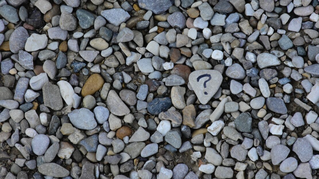 Pebbles on a beach. One pebble has a question mark draw on it.
