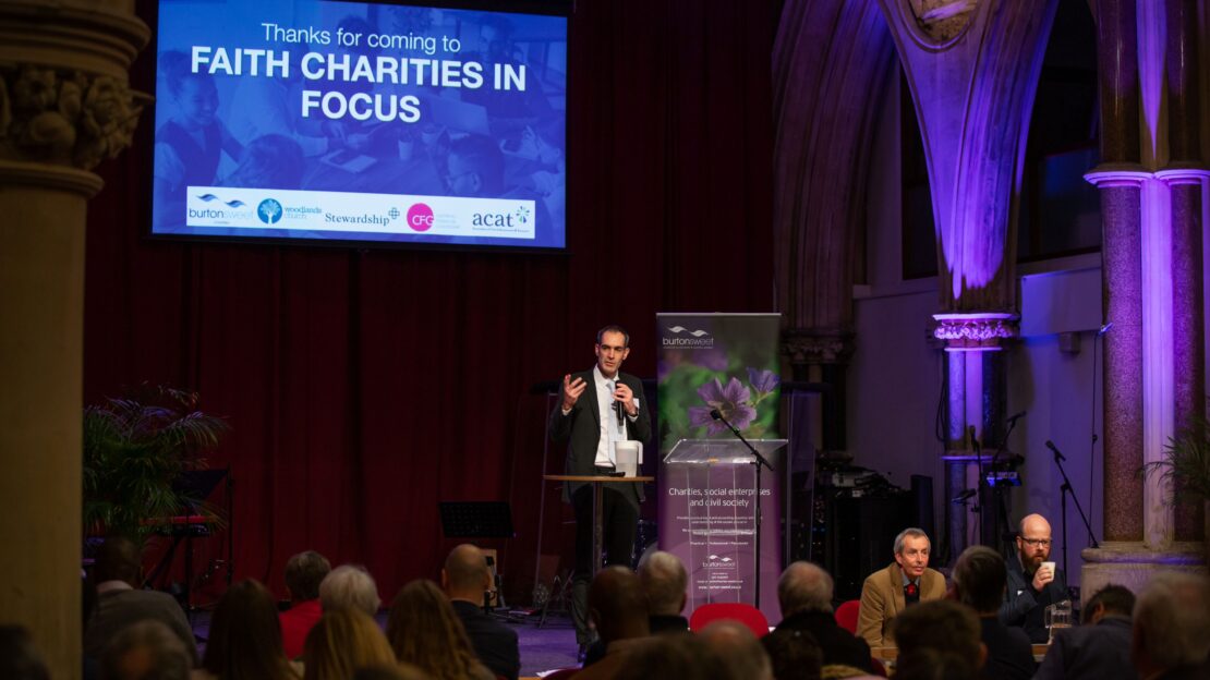 A man in a suit stands on a stage in a beautiful old church. He is wearing a suit and is talking into a microphone. A screen behind him shows a slide that says "Thanks for coming to Faith Charities in Focus".