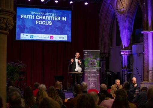 A man in a suit stands on a stage in a beautiful old church. He is wearing a suit and is talking into a microphone. A screen behind him shows a slide that says "Thanks for coming to Faith Charities in Focus".